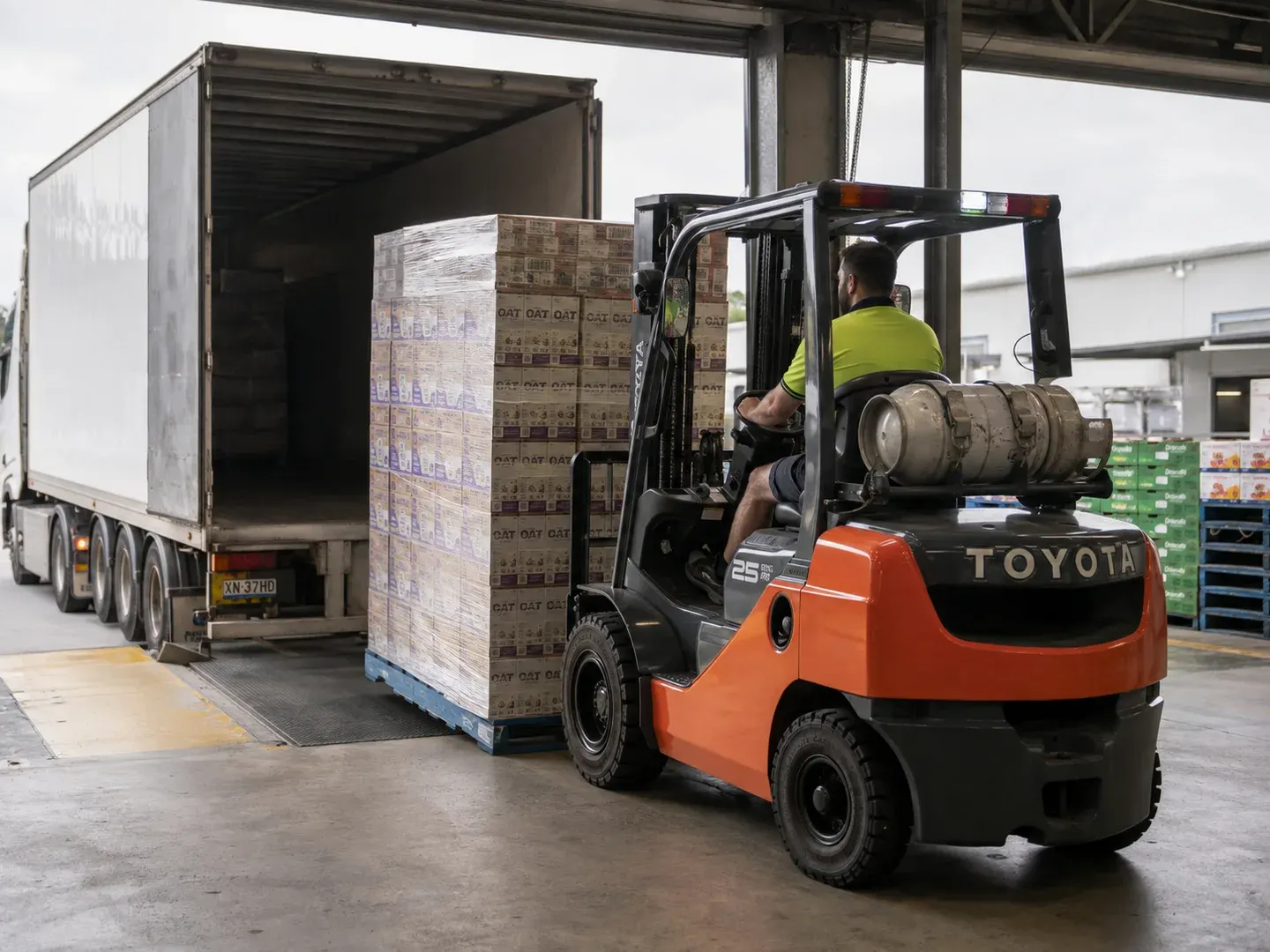 Illustrative scene: An orange 2020 Toyota 8FG25 counterbalance forklift carrying a wrapped pallet of food cartons in an Australian food-wholesaler loading bay, recognisable Toyota branding, accurate forks under pallet, operator safely seated, delivery truck in background. Not the actual customer or their asset.