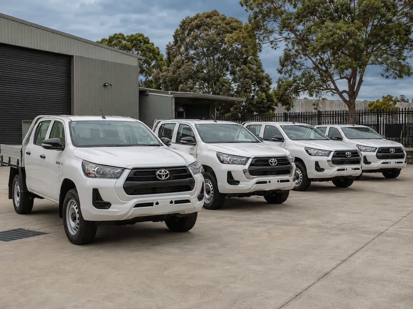 Illustrative scene: Exactly FOUR new white Toyota HiLux double-cab utility vehicles parked in a staggered row at a modest Australian security-services depot. Not the actual customer or their asset.