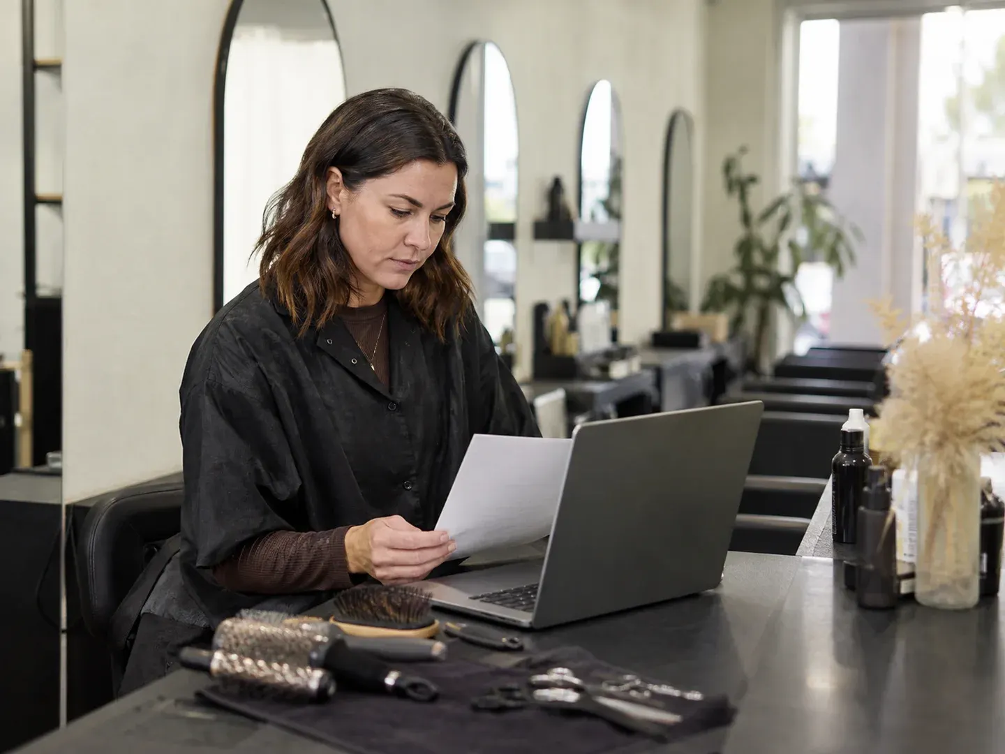 Illustrative scene: An Australian self-employed hairdresser at a compact salon workstation after a workday, looking over a plain document beside a laptop, brushes, scissors and hair products present, empty salon mirrors in background. Not the actual customer or their asset.
