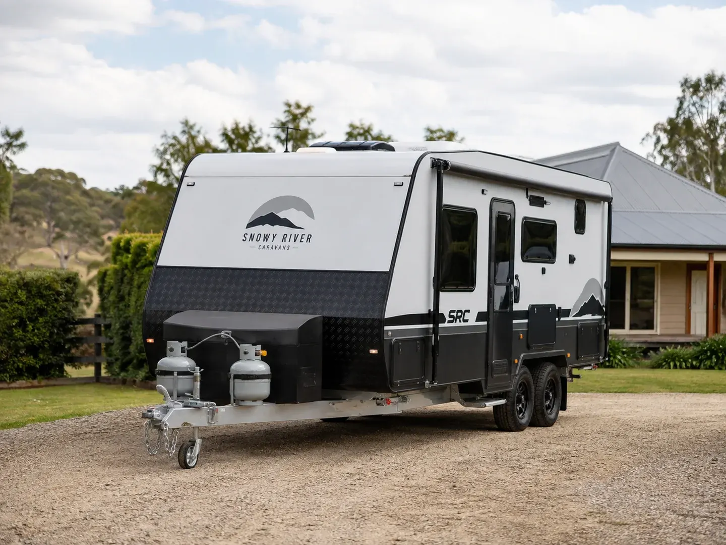Illustrative scene: A 2024 Snowy River SRC caravan parked on a gravel driveway for a private sale in Australia, recognisable Snowy River brand marking and realistic travel caravan shape, complete hitch and caravan visible in three-quarter view. Not the actual customer or their asset.