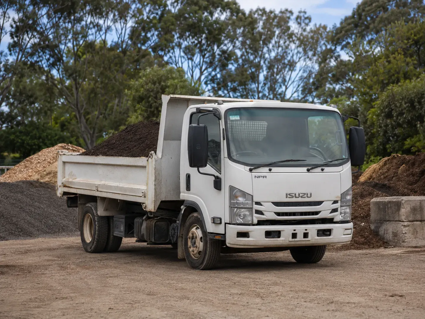 Illustrative scene: A used white 2022 Isuzu NPR tipper truck at an Australian landscaping yard, accurate cab-over NPR cab and Isuzu grille text, tipper tray carrying a modest load of soil, whole truck and tyres visible. Not the actual customer or their asset.