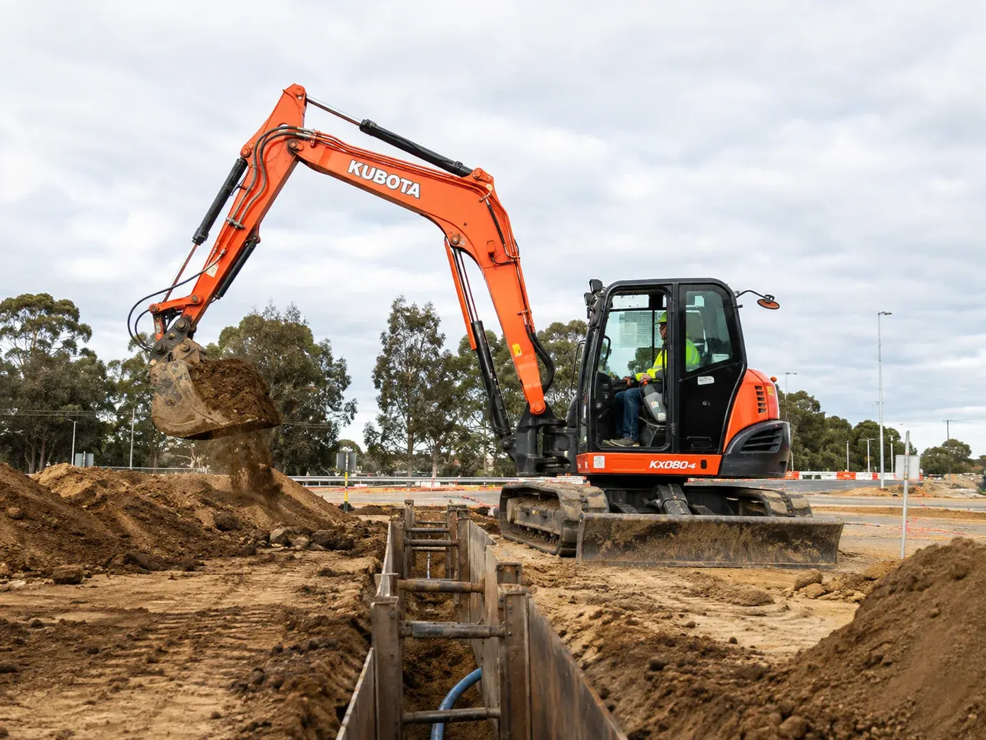 Illustrative scene: A used orange 2019 Kubota KX080-4 excavator working at an Australian civil construction site, recognisable compact 8-tonne Kubota shape and boom branding, steel tracks, bucket moving soil near a service trench. Not the actual customer or their asset.