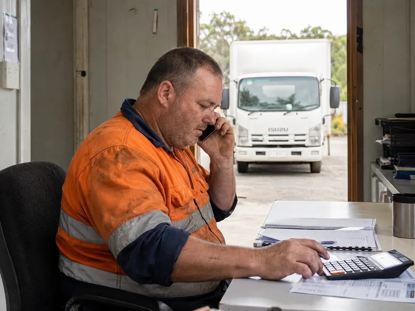 Illustrative business finance scene: A transport business owner reviewing commitments on a phone call, with a white Isuzu delivery truck outside his depot